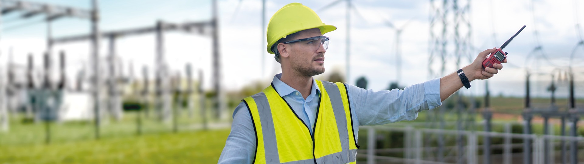 Techniker mit Schutzkleidung steht in einem Umspannwerk, hält ein Tablet und spricht über ein Funkgerät. Im Hintergrund sind Windräder zu sehen